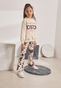 Cream sweatshirt with "BOSTON" text, paired with flared brown and cream speckled pants. White sneakers complete the outfit. Simple background shapes.