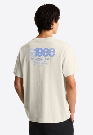 Persona con camiseta color hueso con texto azul “1966 THE NORTH FACE EST. BERKELEY CALIFORNIA” en la espalda, de pie con las manos en los bolsillos.