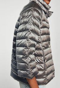 Hopeinen toppatakki korkealla kaulaosalla, jossa on tikattuja paneeleja ja sileä, kiiltävä pinta. Lyhyet hihat ja vetoketjusuljenta mukana.