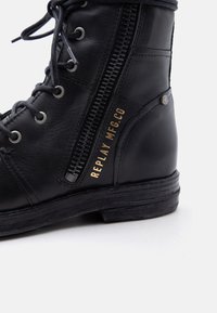 Schwarzer Leder-Schnürstiefel mit seitlichem Reißverschluss und goldenem Schriftzug "REPLAY MFG.CO", robuster Sohle und Metallnieten-Detail nahe der Ferse.