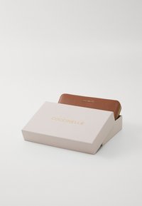 Portefeuille en cuir marron avec finition texturée et marquage doré, à l'intérieur d'une boîte rose pâle, partiellement ouverte pour révéler le portefeuille.