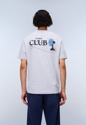 Personne aux cheveux bouclés vue de dos portant un t-shirt gris clair avec le texte "NAPA CLUB" et un dessin de ventilateur de bureau bleu.
