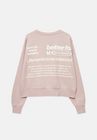 Sweat-shirt rose clair à manches longues avec un texte blanc au dos promouvant l'équilibre, la vie lente, l'harmonie et des attitudes essentielles de vie.