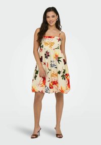 Femme portant une robe crème sans manches, de longueur genou, ornée de motifs floraux et de feuilles colorés, accompagnée de sandales à talons hauts marron.