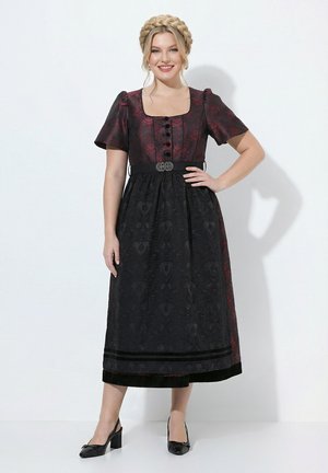 Mujer con el pelo trenzado que lleva un vestido tradicional rojo oscuro y negro con mangas cortas y zapatos negros de tacón, de pie con una mano en la cadera.