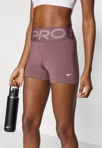 Σορτς Nike Pro σε μωβ απόχρωση, κατασκευασμένα από ελαστικό ύφασμα με φαρδιά ζώνη στη μέση. Αξιοσημείωτο λευκό λογότυπο στο πόδι. Μπουκάλι νερού στο χέρι.