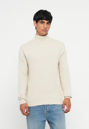 White ribbed turtleneck sweater with long sleeves, made from a textured knit fabric, paired with light blue denim jeans.