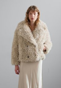 Crème faux fur dubbel-breasted jas met grote revers, gestructureerd oppervlak en donkere knopen; gedragen over een lichtgekleurde satijnen jurk.