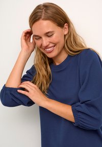 Top a maniche lunghe blu navy con tessuto testurizzato, colletto rotondo e polsini arricciati. Il modello tocca la manica, mostrando la vestibilità comoda.