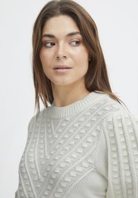Pull gris clair avec des motifs texturés, encolure côtelée et manches bouffantes ; présente un mélange de styles de tricot et de lignes verticales.