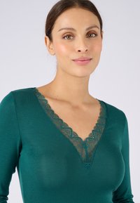 Maglia a maniche lunghe di colore turchese con profondo scollo a V bordato da delicata pizzo. La texture morbida e il design aderente mettono in risalto il décolleté.