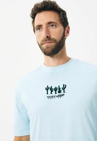 Tricou din bumbac de culoare albastru deschis, cu decolteu rotund, având un design brodat cu un cactus în verde și textul "TERRY URBAN" dedesubt.
