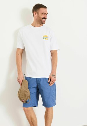Mann lächelt, trägt ein weißes T-Shirt mit Zitronengrafik und blaue Shorts, hält eine beige Baseballkappe und steht vor einem weißen Hintergrund.