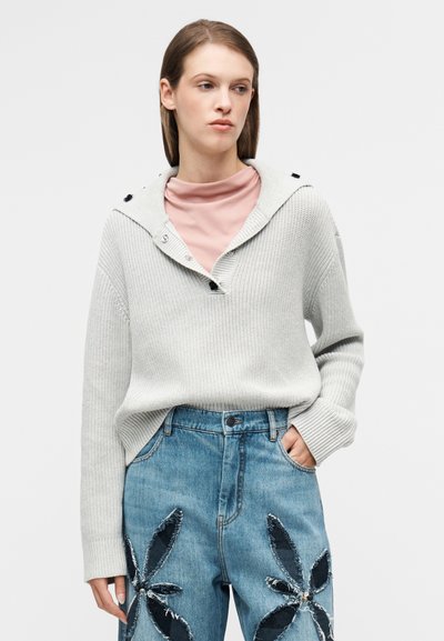 Pull en tricot gris clair avec capuche, doté de boutons et d'une coupe décontractée, associé à un jean bleu orné de patchs en denim fleuris.