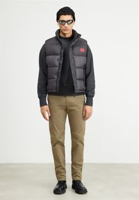 Puffer vest in dark gray with a red logo patch, worn over a black sweatshirt. Paired with khaki pants and black boots.