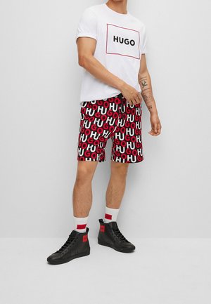 White t-shirt with "HUGO" logo, patterned red and black shorts featuring repeating text, high-top black shoes with red accents, and white socks.