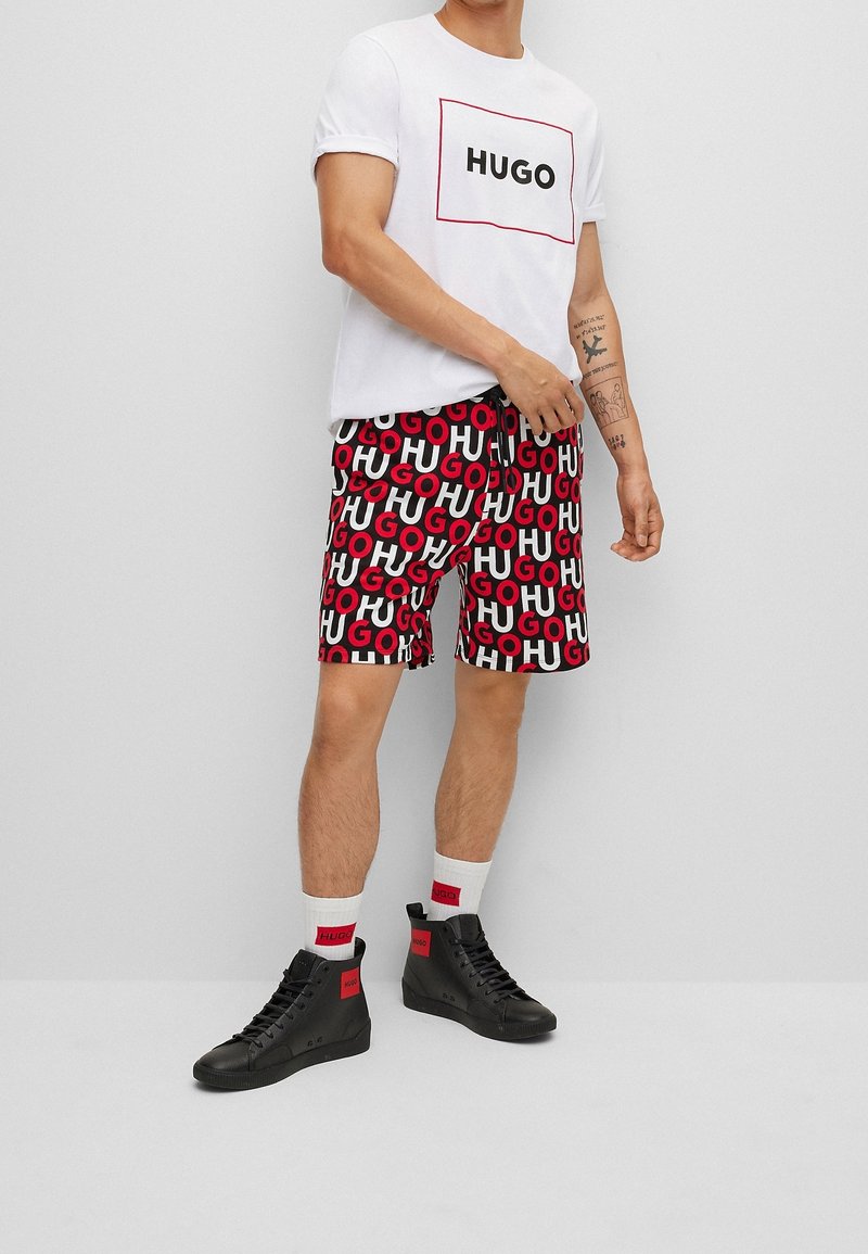 T-shirt blanc avec logo "HUGO", shorts rouges et noirs à motifs avec texte répétitif, chaussures montantes noires avec accents rouges, et chaussettes blanches.