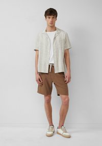 Chemise crème à rayures à manches courtes, t-shirt blanc, shorts marron et baskets avec des accents beiges, présentant une tenue décontractée.