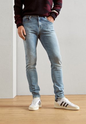 Hombre con jeans ajustados azul claro y zapatillas blancas con rayas negras, de pie sobre suelo de madera frente a una pared lisa.