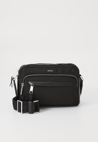 Black fabric shoulder bag with silver hardware, two zippered compartments, a front slip pocket, and an adjustable strap. Features logo detail.