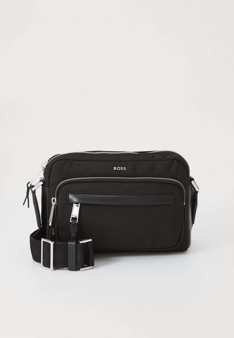 Black fabric shoulder bag with silver hardware, two zippered compartments, a front slip pocket, and an adjustable strap. Features logo detail.