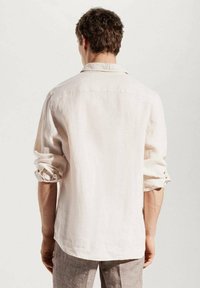 Mężczyzna z brązowymi kręconymi włosami, ubrany w długą, jasnobeżową lnianą koszulę i jasnobrązowe spodnie, stojący tyłem z podwiniętymi rękawami.