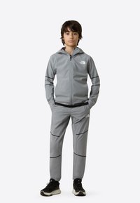 Grå atletisk treningsdress med glidelåsjakke og matchende bukser. Har svarte detaljer og logo. Stoffet virker glatt og tettsittende. Svarte sko.