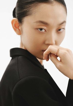 Blazer negro con un cuello estructurado, combinado con una mano en la barbilla que muestra anillos de metal finos. Piel suave y fondo neutro.