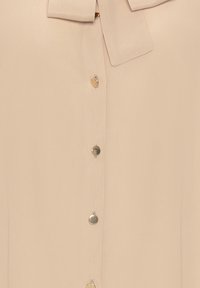 Blusa beige con colletto annodato, texture liscia e bottoni dorati che scendono lungo la parte frontale con una leggera lucentezza.