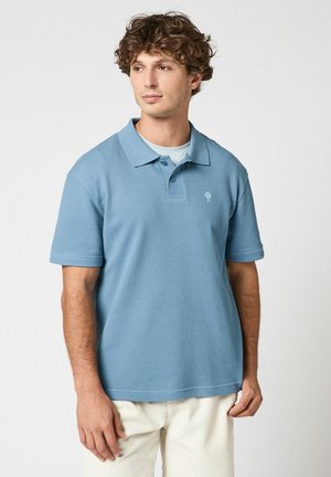 Jeune homme aux cheveux bouclés portant un polo bleu clair texturé et un pantalon blanc cassé, debout devant un fond uni.