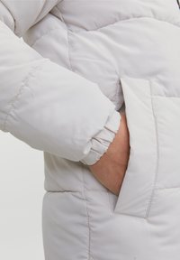 Rękaw jasnoszarej ocieplanej kurtki z elastycznym mankietem; częściowo widoczna boczna kieszeń. Gładki, ocieplany materiał z przeszywanym wzorem pikowania.