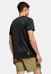 Schwarzes kurzärmliges Sportshirt mit glatter Textur, rundem Ausschnitt und logodetail am Saum. Getragen mit khakifarbenen Shorts.