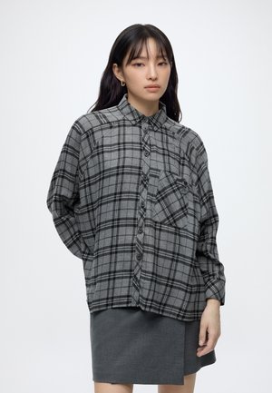 Giovane donna con lunghi capelli scuri indossa una camicia a quadri grigia e nera con bottoni e una gonna grigio scuro su uno sfondo bianco uniforme.