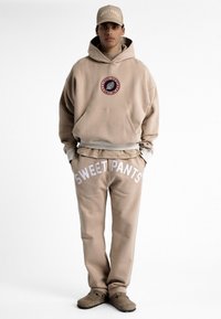 Hoodie en polaire beige avec poche frontale et badge rond ; pantalon de jogging assorti avec "SWEET PANTS" imprimé en blanc. Casquette beige assortie.