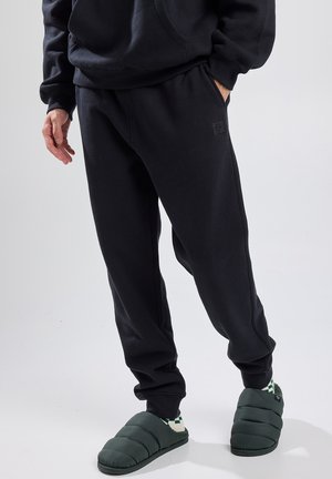 Pantalones de chándal negros de tela suave con puños acanalados, que cuentan con bolsillos laterales y un pequeño parche con el logo. Combinados con zapatillas acolchadas verdes.