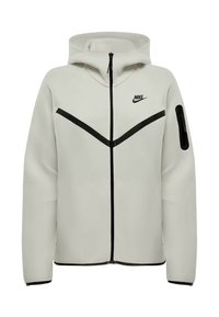 Sudadera con cremallera de Nike en gris claro, con rayas diagonales negras, bolsillo con cremallera en la manga y capucha, presentada de frente sobre un fondo blanco.