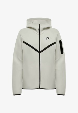 Haut de sweat zippé Nike gris clair avec des rayures diagonales noires, poche zippée sur le bras et capuche, présenté de face sur un fond blanc.
