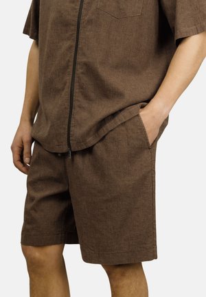 Brune teksturerte shorts og matchende zip-up kortermet topp med hendene i lommer, synlig fra midt på låret til midjen.