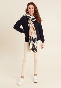 Sciarpa in stampa animale nera e beige appoggiata su un maglione blu, abbinata a pantaloni crema e sneakers bianche, creando un look a strati.