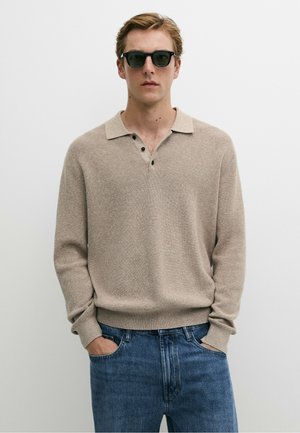 Mann trägt schwarze Sonnenbrille, beige langärmeliger Strick-Poloshirt, blaue Jeans, steht mit Händen in den Taschen vor weißem einfarbigen Hintergrund.