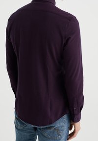 Violetti pitkähiha paita, jossa nappikaulus ja istuva mallitus; siinä on kalvosinnapit ja sileä pinta, yhdistetty vaaleansinisiin farkkuihin.