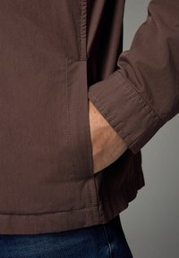 Chaqueta marrón con un acabado texturizado, que presenta un bolsillo lateral y costuras visibles en el puño. Llevada sobre vaqueros.