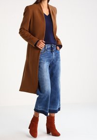 Manteau en laine marron, haut bleu marine à col en V, jean large bleu et bottines rouges. Texture lisse, coupe ajustée et couleurs unies sans motifs.