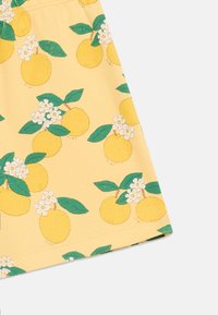Tissu jaune avec un motif imprimé de fruits jaunes, de feuilles vertes, et de petites fleurs blanches, présenté sur fond blanc.