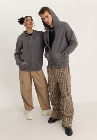 Felpa grigia con zip e cappuccio regolabile, abbinata a pantaloni cargo beige e pantaloni a quadri. Caratteristiche comprendono tasche piatte e trame contrastanti.
