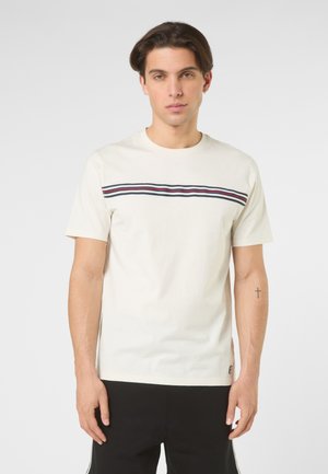 Junger Mann trägt ein weißes kurzärmeliges T-Shirt mit horizontalen roten, weißen und schwarzen Streifen über der Brust, kombiniert mit einer schwarzen Hose.