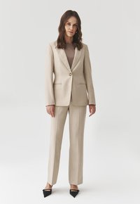 Beige přizpůsobený blazer s jedním knoflíkem, doplněný o odpovídající kalhoty. Hladká tkanina, ostré klopy a decentní elegance. Černé špičaté boty.
