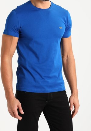 Homme portant un t-shirt bleu ajusté à manches courtes avec un petit logo sur la poitrine et un pantalon noir sur fond blanc.