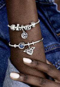 Silberne Charm-Armbänder an einem Handgelenk, mit einer Mischung aus Charms, darunter ein blauer Stein und ein Herz mit Flügeln. Glatte, polierte Oberflächen.