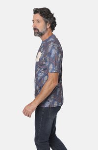 Camisa de manga corta azul con un patrón de hojas de palmera en crema y marrón. Incluye un pequeño bolsillo en el pecho con un detalle de logo.